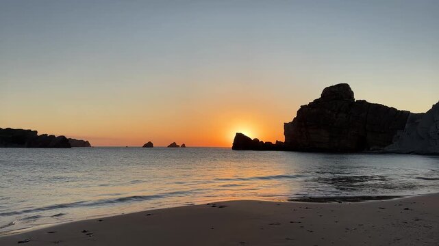 Golden sunset as the sun slowly sets behind a rocky islet, illuminating a calm rocky cove with gentle waves and warm evening light.
