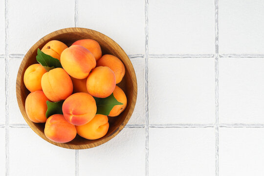 Ripe apricots with green leaves arranged in wooden bowl. Fresh seasonal harvest, summer fruit on rustic kitchen, white tile background with copy space healthy eating, dessert, food ingredient