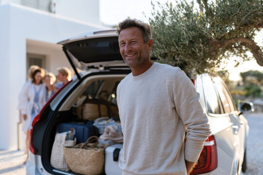 A happy man stands beside a car packed for a vacation, showcasing excitement and adventure as family members gather in the background, ready for a memorable journey.