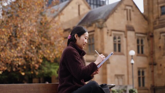 An Asian woman sits on an outdoor bench, focused on drawing and writing on a digital tablet with a stylus. A historic university building and autumn trees complete this study scene