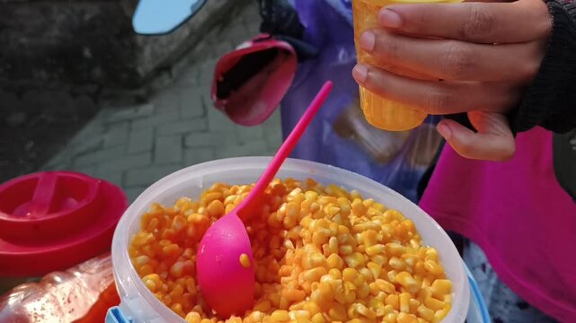 Street food vendor scooping sweet corn kernels into a plastic cup.