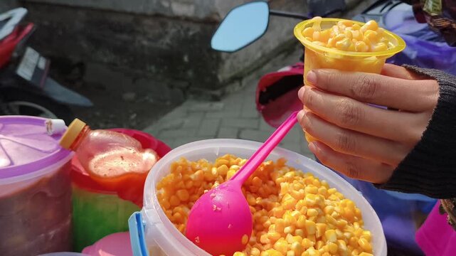 Street food vendor scooping sweet corn kernels into a plastic cup.