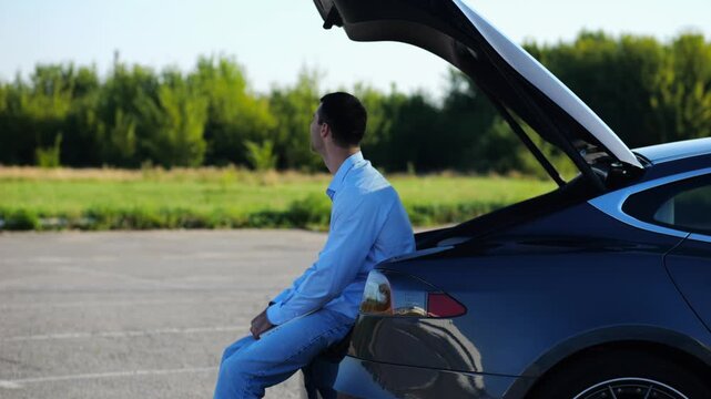 Successful businessman in formal wear finished work on laptop sitting in electric car trunk. Young handsome entrepreneur resting after online job in modern automobile. Concept of freelance work