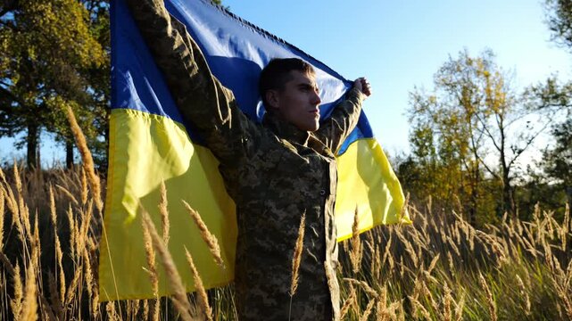 Military man in uniform stands with raised over head flag of Ukraine at countryside. Male ukrainian army soldier with lifted national banner in honor of victory against russian aggression. End of war