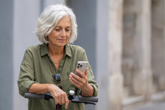 Mature gray haired woman leaning on electric scooter and checking mobile phone outdoors