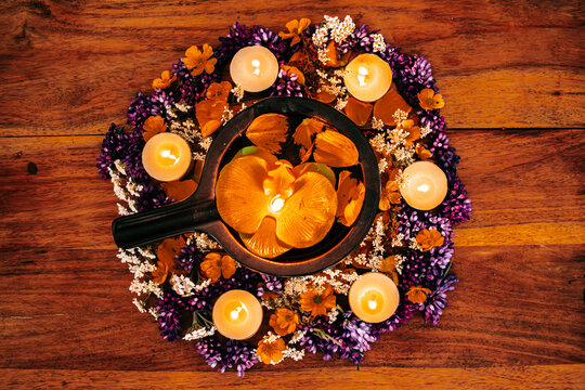 Overhead view of a black metal bowl with a floating golden orchid, surrounded by a circle of tea light candles and mixed purple and orange flowers on a rustic wooden table.