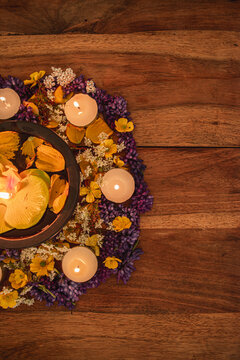 Overhead view of a black metal bowl with a floating golden orchid, surrounded by a circle of tea light candles and mixed purple and orange flowers on a rustic wooden table.