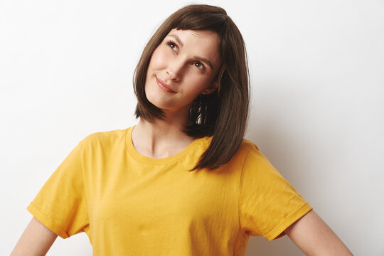 A young brunette woman wearing a yellow shirt stands in a bright studio with a clean white background, posing with a slight head tilt and a calm thoughtful expression.