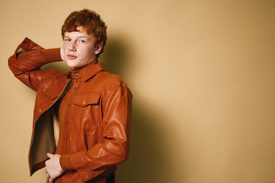 Young man with red hair wearing brown leather jacket posing with hand behind head against neutral background for fashion and style portrait.