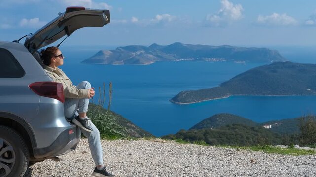 Tourist enjoying breathtaking seascape from car trunk. Woman sitting on open car trunk, overlooking scenic coastal landscape with turquoise waters and distant islands, embodying freedom and wanderlust