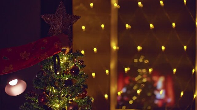 Person Placing a Glittering Star on Top of a Decorated Christmas Tree With Warm Fairy Lights and Fireplace Reflection in the Window, Cozy Holiday Tradition