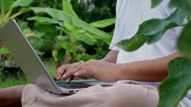 Young professional remote working, laptop on lap while sitting in peaceful garden, free range chickens, relaxed outdoor workspace promoting work life balance, flexible lifestyle, digital productivity