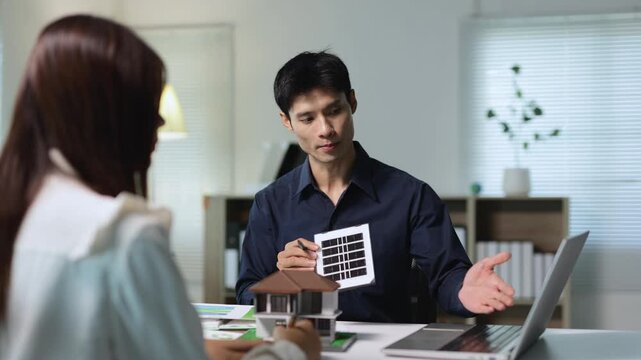 Asian solar energy consultant explaining photovoltaic panels for a new home to a female client in a modern office