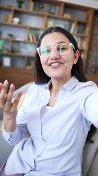 POV Vertical Smiling woman talking and waving during video call indoors sitting on sofa in living room at home. Happy female showing friendly communication looking at camera, has online chat with frie