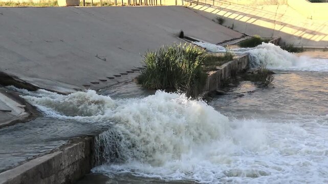 Turbulent water rushing out of industrial concrete spillway channels into a rushing river dynamic splashing stream from irrigation drainage system.