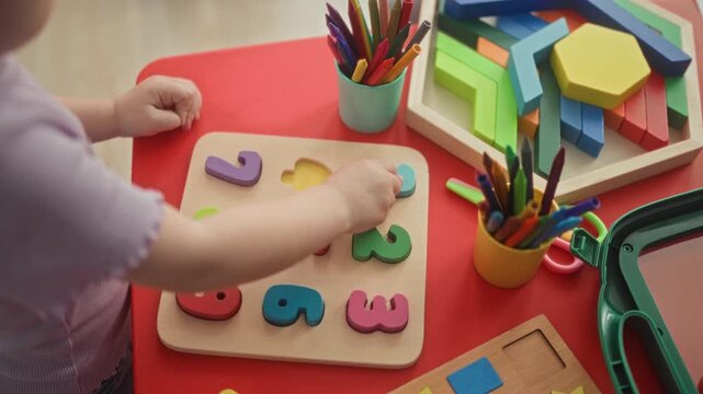 Girl toddler places a wooden number puzzle piece with her hand on a red table in a daycare building among colorful blocks and pencil cups; curiosity learning play.