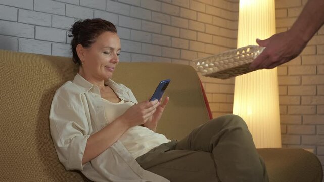 Woman surrendering smartphone during digital detox at home. Smiling woman sitting on a couch gives her smartphone to a man, taking a break from technology and social media for a digital detox
