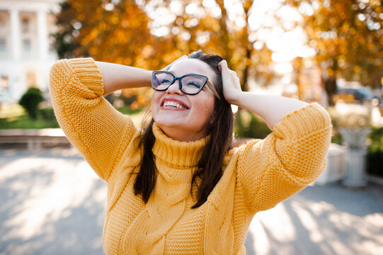 Smiling stylish woman 35-39 year old wearing knitted sweater and eye glasses in city street over yellow leaves. Autumn season.