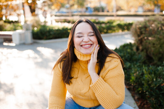 Smiling stylish woman 35-39 year old with eyes closed wearing knitted sweater in city street over yellow leaves. Autumn season.