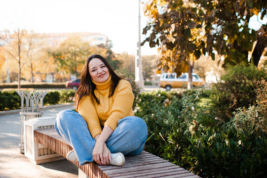 Smiling stylish woman 35-39 year old wearing knitted sweater sitting on bench at city street over yellow leaves. Autumn season.