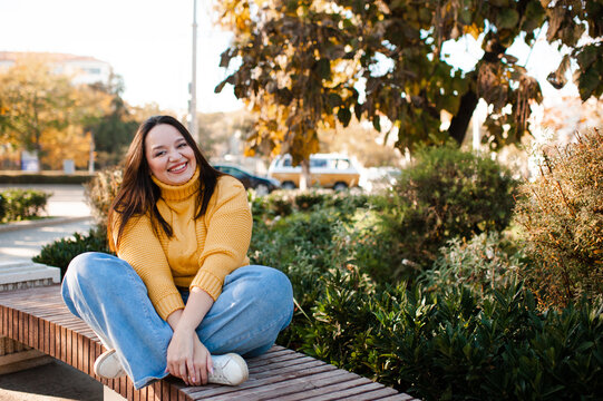 Smiling stylish woman 35-39 year old wearing knitted sweater sitting on bench at city street over yellow leaves. Autumn season.