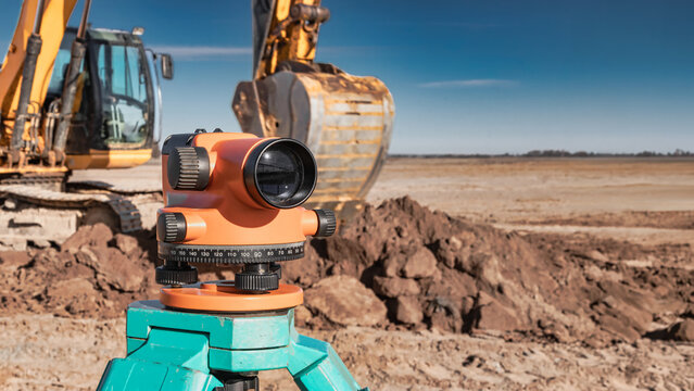 Workers use a geodetic level to measure land at a construction site. Heavy machinery operates nearby on a wide expanse of ground under a blue sky