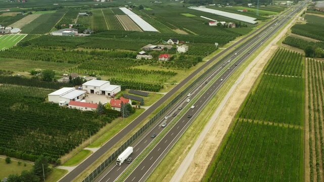 Automobiles and freight trucks drive on a long highway through countryside. Active vehicle traffic moves along an asphalt freeway passing organic agricultural farms on a summer day.