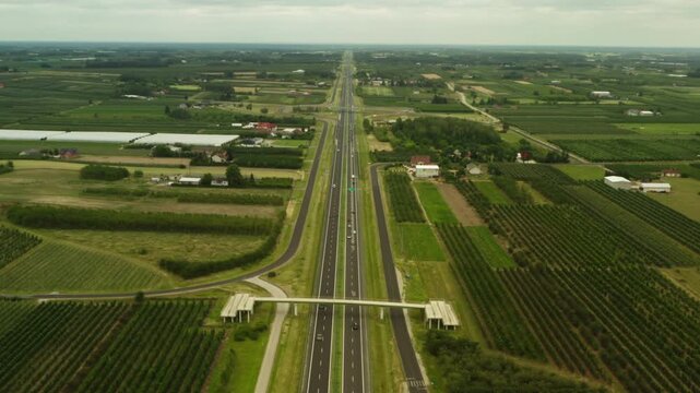 Vehicles drive on a straight highway passing through agricultural farmlands. Active transit traffic moves along a multi-lane asphalt freeway near green rural orchards on a summer day.