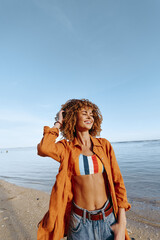 Young woman with curly hair enjoys a sunny day on the beach wearing a colorful swimsuit and orange shirt.