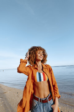 Young woman with curly hair enjoys a sunny day on the beach wearing a colorful swimsuit and orange shirt.