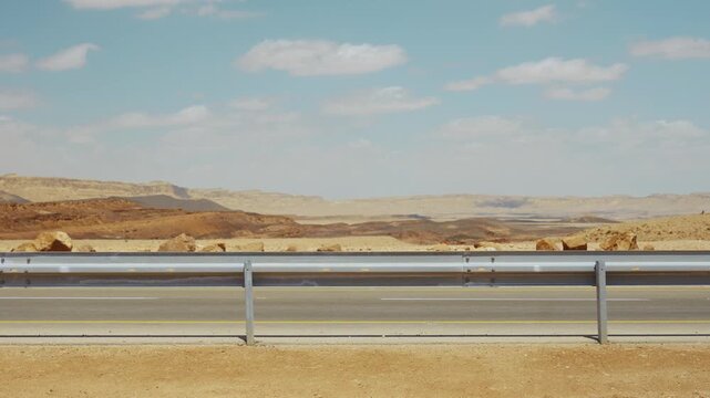 Guardrails line a desert road under a clear blue sky. Distant rocky hills stretch across the horizon. White van speeds past the guardrail. Ramon Crater in Israel Negev region. Warm day outdoors