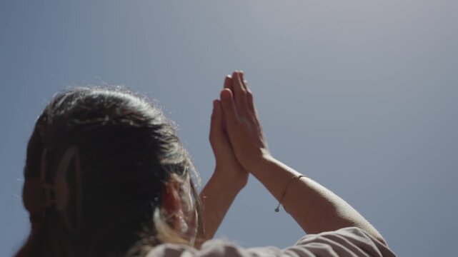 Female with hands clasped praying towards blue sky sunlight. Young religious woman raising her hands upwards in forgiveness or empowerment. Faith or spirituality. Meditation and peacefulness outdoors