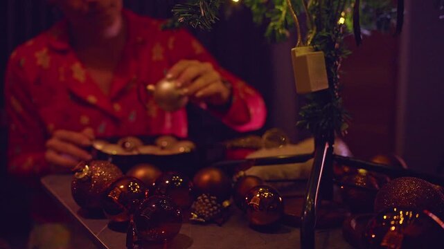 Christmas Ornaments Spread Across a Table as a Woman Prepares Decorations for a Festive Tree, Cozy Holiday Atmosphere and Warm Fireplace
