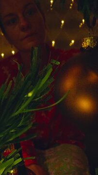 Vertical video. Creative Point of View From Inside a Christmas Tree as a Woman Carefully Places a Holiday Ornament Among Glowing Branches and Warm Fairy Lights