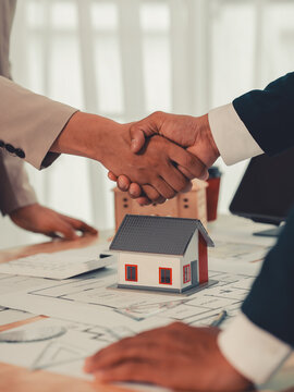 A real estate agent and an architect shake hands over a house model and blueprints on a desk to celebrate a successful deal; close-up shot.