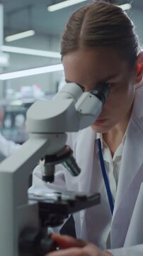 Vertical Screen. Caucasian Woman Scientist Peers Through Optical Microscope Lens, Adjusting Focus Knob While Analyzing Specimen in Medical Laboratory. Researcher Advances Drug Development.