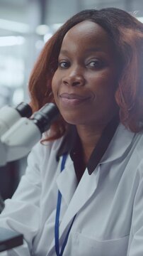 Vertical Screen. Smiling Black Female Scientist Sits Beside Optical Microscope, Looking at Camera in Portrait Pose Within Modern Medical Laboratory. Researcher Drives Pharmaceutical Innovation.