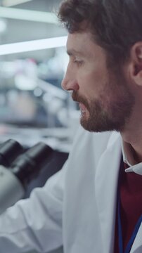 Vertical Screen. Focused Caucasian Male Scientist Peers Into Optical Microscope, Examining Specimen Sample in Modern Medical Research Laboratory. Physician Conducts Analysis for Drug Development.