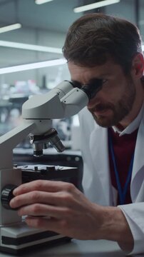 Vertical Screen. Caucasian Male Scientist Adjusts Focus Knob While Looking Through Optical Microscope, Studying Specimen Slide in Modern Medical Research Lab. Physician Advances Drug Development.