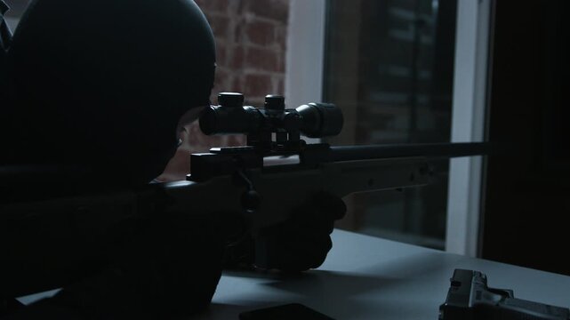 Cinematic footage of a masked sniper aiming a scoped rifle out of a window in a dark room with brick walls.