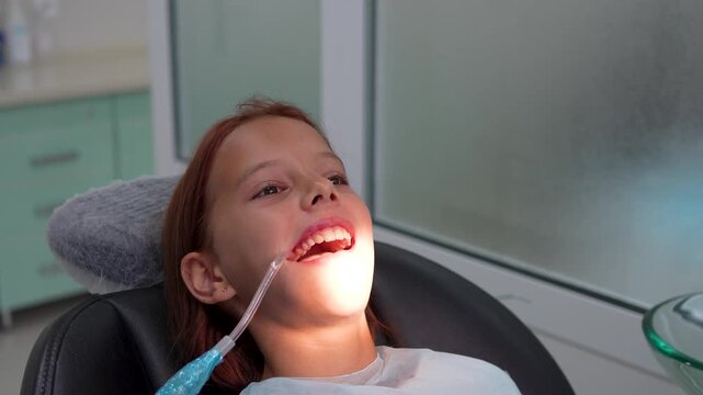 Little girl with open mouth using saliva ejector in dental chair close up.