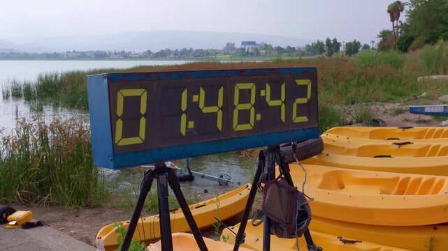 Digital sports scoreboard timer clock on a tripod showing race time during a triathlon and kayaking competition by the lake shore. Perfect stock video for sporting events and tournament concept.