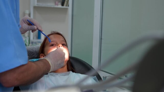 Little girl with open mouth and saliva ejector sitting in dental chair under lamp close up.