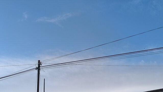 Blue sky with soft clouds above power lines. Wooden pole supports multiple cables stretching across frame. Daylight scene shows urban infrastructure and open atmosphere. Background provides clear spac