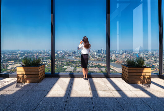 A business woman talks on her phone looking out of her office window high above the city skyline of London, England