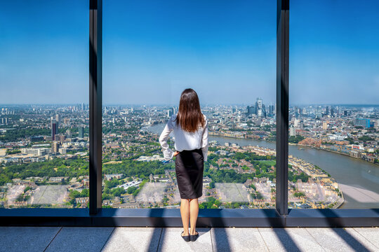 Rear view of a business woman looking out of her office window high above the urban skyline of the City of London, England
