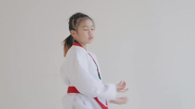 A child shows karate moves in a training room with white walls. The child wears a white uniform with a red belt. The setting is bright and simple, focusing on the training.