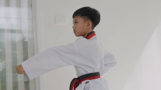 A girl practices her martial arts skills at home. She carefully ties her red belt around her waist while wearing a traditional uniform. The room is well lit and simple.