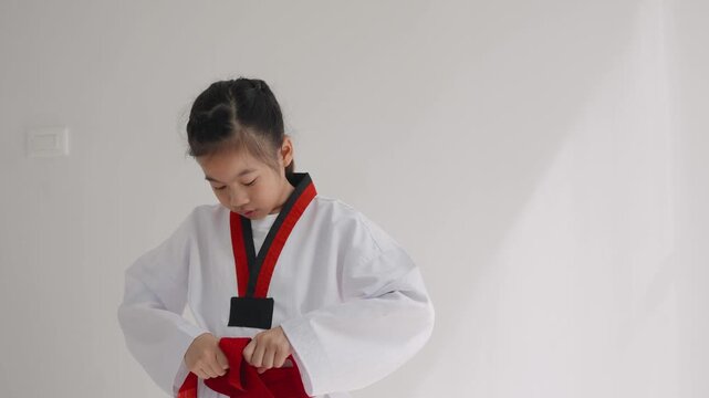 A girl practices her martial arts skills at home. She carefully ties her red belt around her waist while wearing a traditional uniform. The room is well lit and simple.
