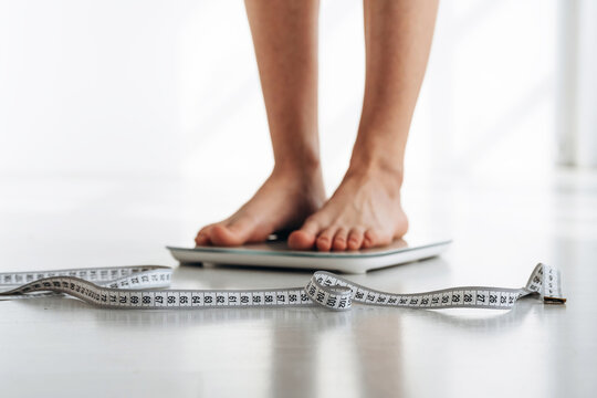 Barefoot individual standing on a digital scale with a measuring tape lying on the floor, bright indoor setting with natural light illuminating the scene
